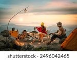 Campfire in the midst of nature. There is an Asian couple making coffee and playing music together while setting up a tent on a mountaintop with a view of nature in Thailand while the sun is setting.