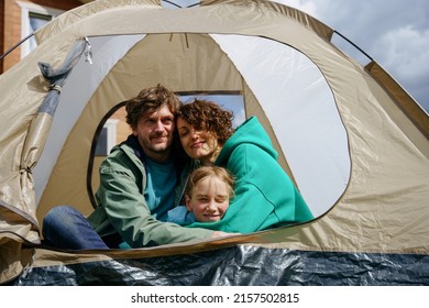 Camping In The Backyard. A Family Is Sitting With A Tourist Tent, Hugging And Looking At The Camera In The Backyard