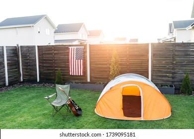 Camping in the backyard. An orange tent, folding chair and guitar on the lawn, american flag hang on fence. Leisure time, independents day