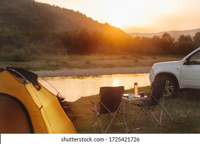 Camping At Beach Of Mountain River. Suv Car. Yellow Tent. Camp Table And Chairs