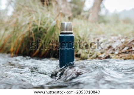 Camping blue thermos standing in stream of river, close-up of vacuum flask covered with water drops in nature.