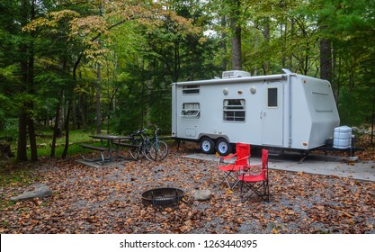 Camping In Camper Trailer In Wooded Park Camp Site With Chairs Set Up Around Fire Pit, And Bicycles Leaning Against Picnic Table.