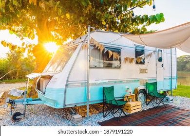 Camping chairs placed outside cozy retro travel trailer Caravan under tree before sunset near the river in peaceful countryside. Outdoor and Recreational Vehicles Theme. Travel Industry.