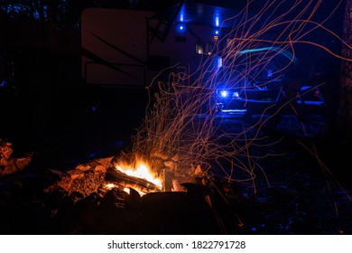 A Camping Fire With Embers Flowing Around, Blue Lights From The Camper In The Back Drop