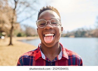 Camping, funny face and portrait of child in nature for playing, joke or hiking vacation. Lake, African or happy black boy with tongue out for outdoor, travel, summer break or adventure in Nigeria - Powered by Shutterstock