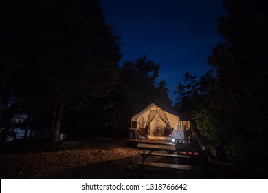 Camping Or Glamping Tent Site With Camp Fire Under The Blue Twilight Sky With An Illuminated Tent And Trees.