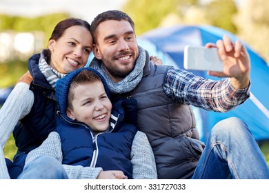 Camping, Hike, Technology And People Concept - Happy Family With Smartphone Taking Selfie At Campsite