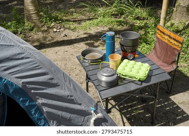 Camping kitchen setup with cookware, kettle, stove and eggs on a portable table beside a tent in a forest campsite.