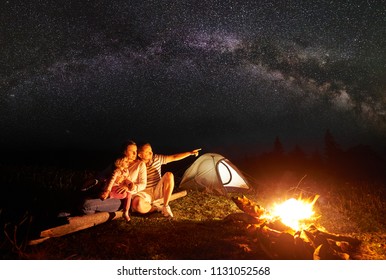 Camping In Mountains At Night. Tourist Family Resting In Front Of Illuminated Tent And Burning Bonfire. Woman Holding In Arms Small Girl, Man Pointing At Bright Stars In Dark Sky And Milky Way