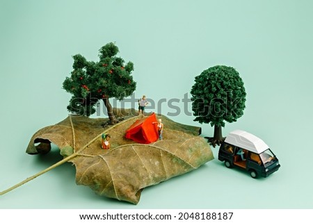 Camping in nature on a dry autumn leaf with a small tent, miniature people, two trees and a camping van on pastel green background. Creative camping or fall outdoor activity concept.