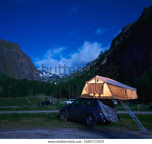 Camping night scene with silver car, majestic\
hills on background. Beautiful view of blue evening sky over high\
mountains and SUV with rooftop tent. Concept of travelling, camping\
and mountain hiking.