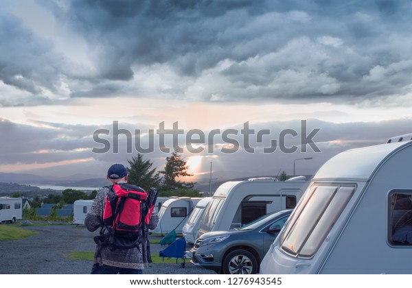 Camping parking\
with lot of caravans, trailers, cars, campers. mature man walking\
with red rucksack, view from\
back