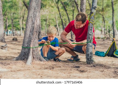 Camping People Outdoor Lifestyle Tourists In Summer Forest Near Lazur Sea. Blond Boy Son With Father Study Survival Techniques, Practice Methods Of Tying Rope Knots. Natural Children Education