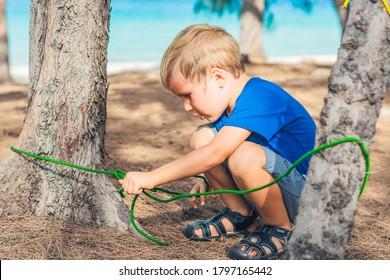 Camping People Outdoor Lifestyle Tourists In Summer Forest Near Lazur Sea. Blond Serious Boy In Blue T-shirt Study Survival Techniques, Practice Methods Of Tying Rope Knots. Natural Children Education