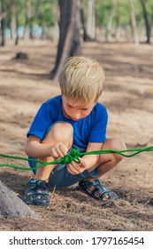 Camping People Outdoor Lifestyle Tourists In Summer Forest Near Lazur Sea. Blond Serious Boy In Blue T-shirt Study Survival Techniques, Practice Methods Of Tying Rope Knots. Natural Children Education