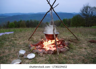 The Camping Pot On The Open Fire In The Nature With The Blue Mountains In The Background During Sunset. The Photo Of The Campfire With The Kettle On The Horizon Of The Park.