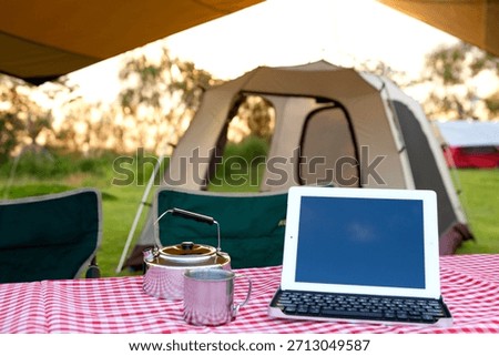 Camping setup with a tent, kettle, and tablet in a green area during sunset with a relaxed outdoor vibe
