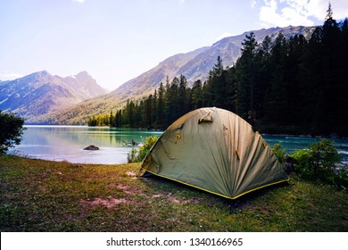 Camping Tent In Campground At National Park. Tourists Camped In The Woods On The Shore Of The Lake On The Hillside. View Of Tent On Meadow In Forest. Camping Background