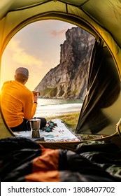 Camping with tent in Lofoten islands, Northern Norway, Kvalvika beach, during sunset. Person sitting in front of a tent holding a hot drink mug.