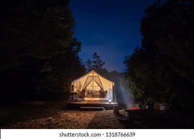 Camping Tent Site With Camp Fire Under The Blue Night Sky With An Illuminated Tent And Trees In The Background