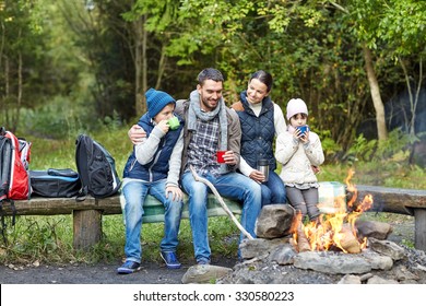 Camping, Travel, Tourism, Hike And People Concept - Happy Family Sitting On Bench And Drinking Hot Tea From Cups At Camp Fire In Woods