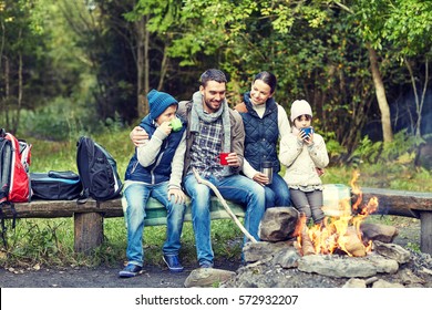 Camping, Travel, Tourism, Hike And People Concept - Happy Family Sitting On Bench And Drinking Hot Tea From Cups At Camp Fire In Woods
