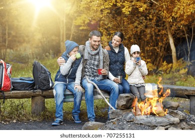 kamperen, reizen, toerisme en mensen concept - gelukkige familie zittend op de bank en het drinken van hete thee uit kopjes bij kampvuur in de herfst bos