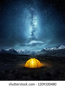 Camping in the wilderness. A pitched tent under the glowing  night sky stars of the milky way with snowy mountains in the background. Nature landscape 