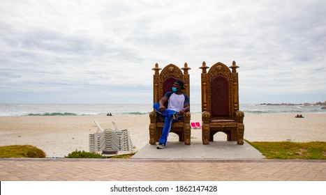 Camps Bay-  Cape Town, South Africa - 11-23-20

Man Sitting On One Of The Royal Thrones Of Camps Bay Beach. Wearing Blue Face Mask. Beach In Background.