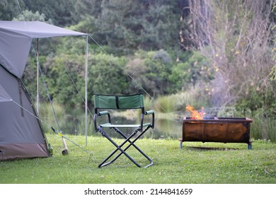 Campsite Along River With Campfire, Canvas Tent And Camping Chair. Outdoor Leisure Campground In Nature.