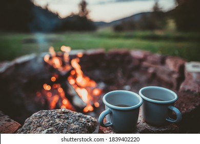 Campsite With Fire Pit and Two Tin Cups with hot tea. Burning Campfire with mountain landscape with evening sunset sky over the forest and hills.