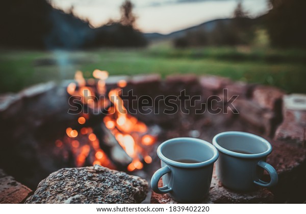 Campsite With Fire Pit and Two Tin Cups with hot\
tea. Burning Campfire with mountain landscape with evening sunset\
sky over the forest and\
hills.