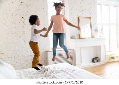 I Can Fly. Naughty Laughing African Children Brother And Sister Jumping On Bed At Home. Happy Black Kids School Age Girl And Preschool Boy Having Fun Playing Active Game In Bedroom At New Big House.