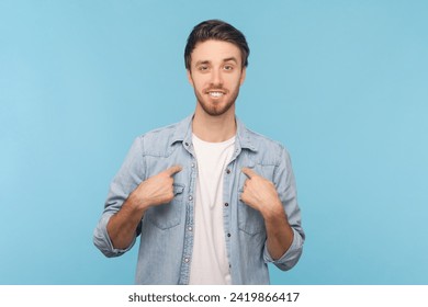 I can handle it. Assertive determined man points at himself, feels proud and brags about own achievements, proposes own help, wearing denim shirt. Indoor shot isolated on blue background.