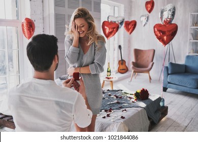 Can not believe this is happening! Surprised young woman covering face with hand and smiling while her boyfriend proposing her in the bedroom