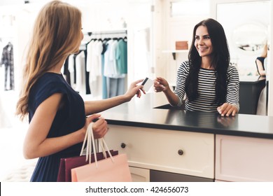 Can I Pay With My Card? Beautiful Young Woman With Shopping Bags Giving Her Credit Card To Seller While Standing At The Clothing Store