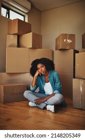 Can Someone Help Me. Portrait Of A Unhappy Looking Young Woman Seated On The Ground While Being Surrounded By Cardboard Boxes On Moving Day Inside At Home.