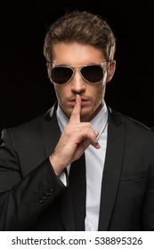 Can You Keep A Secret? Vertical Studio Shot Of A Handsome Young Secret Service Special Agent Wearing A Suit And Sunglasses Shushing With His Finger To His Lips Asking For Silence On Black Background