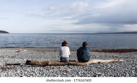 Gaspé, Canada - August 11 2020 : Couple Siting On A Tree Trunk In Front Of The Sea (Gaspe, Quebec) During A Cloudy Day