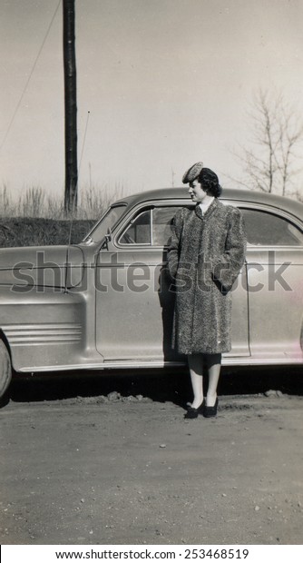 CANADA - CIRCA 1940s: Reproduction of an antique\
photo shows young woman in a fur coat and astrakhan hat posing near\
a luxury car