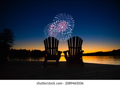 Canada day fireworks on a lake in Muskoka, Ontario Canada. On the wooden dock two Adirondack chairs are facing the sunset orange hues 