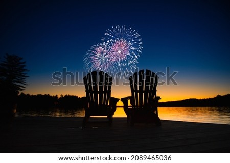 Canada day fireworks on a lake in Muskoka, Ontario Canada. On the wooden dock two Adirondack chairs are facing the sunset orange hues 