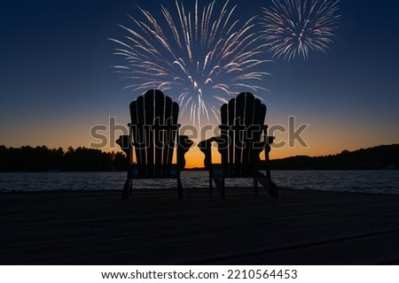 Canada Day fireworks over two Adirondack chairs on the wooden dock in Muskoka, Ontario Canada, are facing the sunset orange hues while facing the calm water.