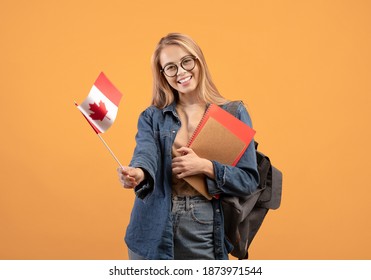 Canada Day, Holiday, Confederation Anniversary. Cheerful Teen Blonde Female In Glasses, With Backpack And Books Waving Small Flag, Isolated On Orange Background, Copy Space, Studio Shot