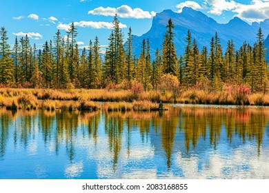 Canada. Indian summer in the Rocky Mountains. The smooth water of Lake Vermillon reflects the snow-white clouds. Yellow and orange foliage of the autumn forest.