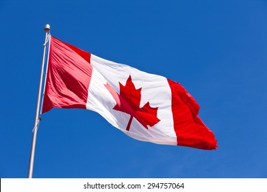 Canadian Flag Flying In Light Breeze On Top Of Metal Pole Against Blue Sky