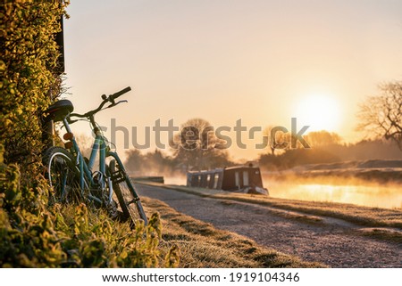 Canal boat with mountain bike left leaning against hedge row early morning sunrise dawn with golden light in sky on the River Trent and mist rising in Nottingham Nottinghamshire