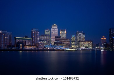 Canary Wharf at night. Illuminated financial district skyline in London, UK. Office windows lights on the skyscrapers of the business district at dusk. Reflection on River Thames.