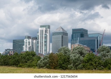 Canary Wharf Skyscrapers - London Financial Hub - View From The Park Including Green Grass Meadow, Bushes And Dark Storm Clouds - London, UK.