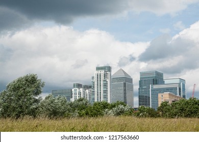 Canary Wharf Skyscrapers - London Financial Hub - View From The Park Including Green Grass Meadow, Bushes And Dark Storm Clouds - London, UK.